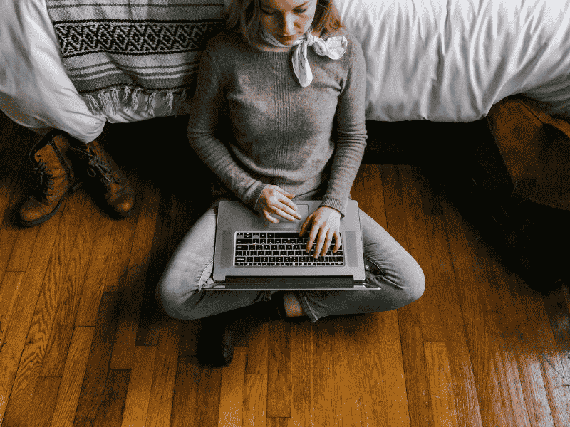 woman wearing a sweater, jeans and socks sits on bedroom floor browsing on her computer.