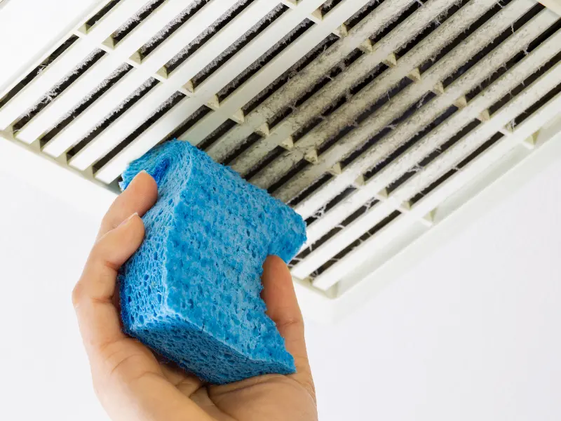 cleaning your air duct covers helps keep down allergens and lets your HVAC run more efficiently. Photo shows a woman holding a blue sponge and wiping dust off a vent register.