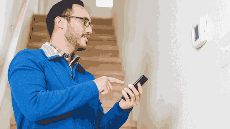 man wearing a blue sweater looks at wall thermostat while adjusting winter thermostat settings on his phone