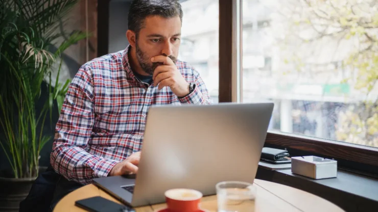business owner wearing a plaid shirt looks thoughtful as he shops for electricty plans on his laptop
